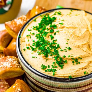 Close up head on shot of a bowl of thick tofu beer cheese topped with minced green parsley and surrounded by mini soft pretzel bites
