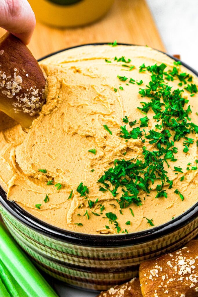Head on close up shot of a bowl of thick vegan beer cheese with a pretzel bite dipping into one side and minced parsley on the other side