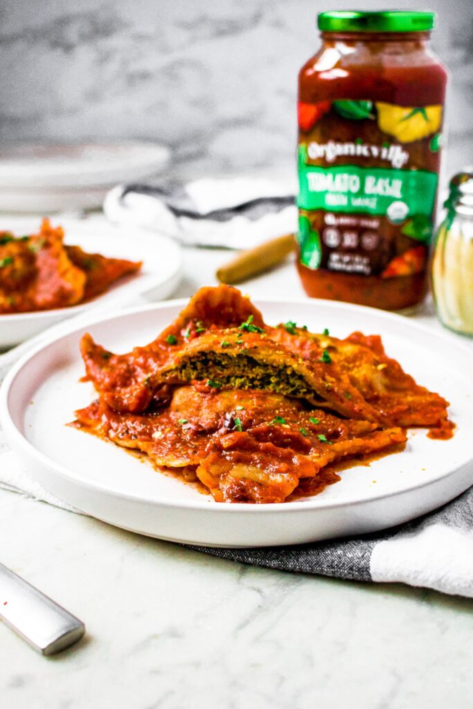 Head on shot of vegan ravioli stacked on a plate and coated in tomato sauce. The top ravioli is cut in half so you can see the dairy free spinach ricotta filling.