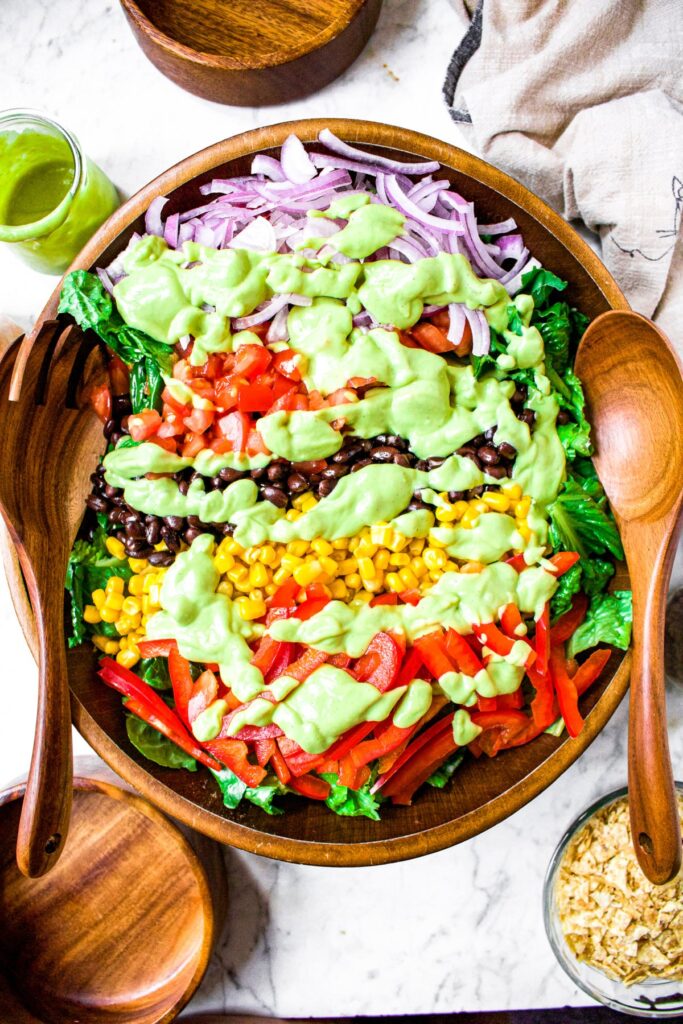 Overhead shot of a large bamboo salad bowl with greens, corn, red peppers, tomato, red onion, and black beans drizzled with creamy avocado dressing. There are bamboo salad tongs on either side of the bowl.