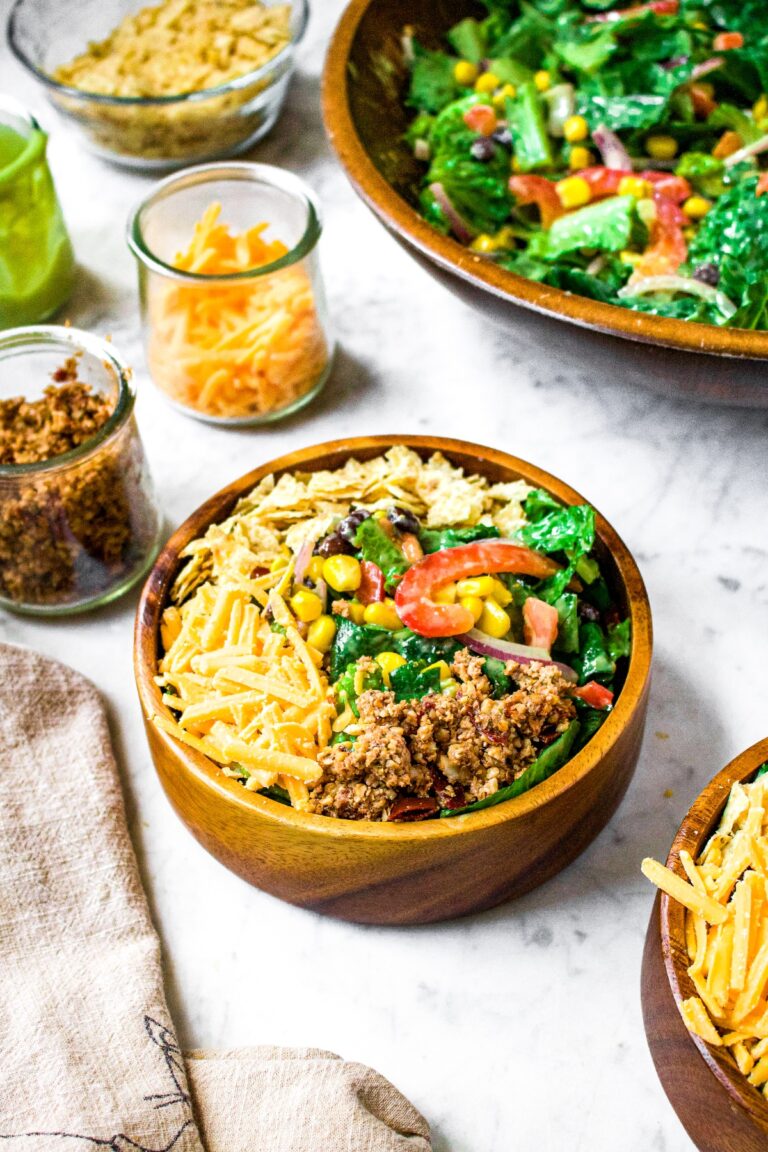 Overhead shot of a small bamboo salad bowl filled with a green southwest salad topped with dairy free cheddar, vegan chorizo, and tortilla chips. There is a larger salad bowl in the background along with small clear jars of shredded cheddar, walnut chorizo, and avocado dressing.