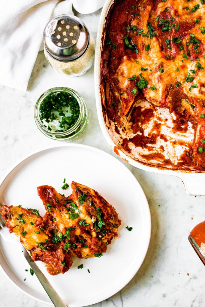 In the bottom left corner, there is a round plate with three vegan stuffed shells and a fork. In the top right corner, there is the full baking dish of stuffed shells.
