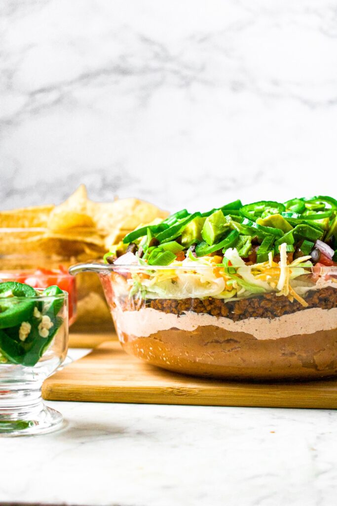 Head on photo of a glass container with meatless taco dip layered in it. There is a jar of sliced jalapenos in front and to the left of the container