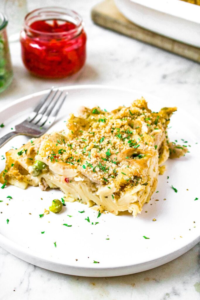 Close up head on shot of a triangular slice of plant based tuna noodle casserole. There is a fork and a jar of pimientos behind the plate