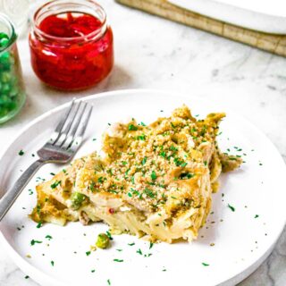 Close up head on shot of a triangular slice of plant based tuna noodle casserole. There is a fork and a jar of pimientos and peas behind the plate