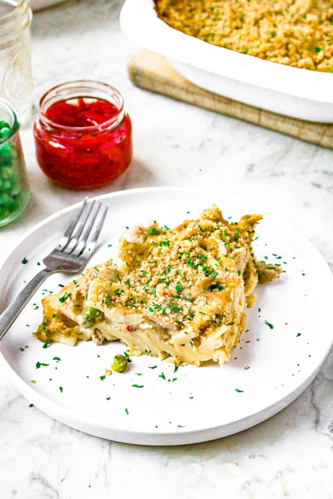 Close up head on shot of a triangular slice of plant based tuna noodle casserole. There is a fork and a jar of pimientos and peas behind the plate