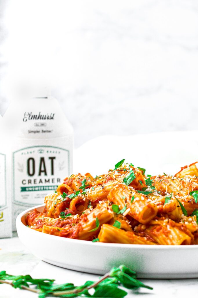 Head on shot of a plate piled high with vegan vodka pasta sprinkled with fresh oregano. There is a sprig of oregano in front of the plate, and a carton of Elmhurst unsweetened oat creamer behind it.