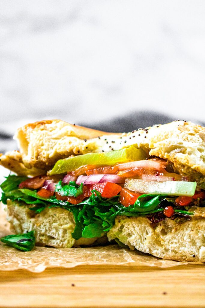 Close up head on shot of a garden vegetable bagel sandwich sliced in half to display the layers of veggie fillings: spinach, tomato, cucumber, red onion, pickles, and roasted red peppers.
