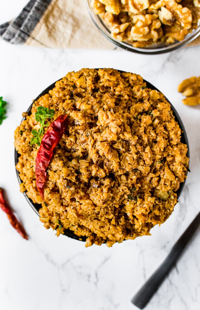 A close-up photo of vegan walnut taco meat in a green and brown striped bowl. There is a single dried red chili on top along with a sprig of oregano. A ramekin of raw walnuts is in the background.