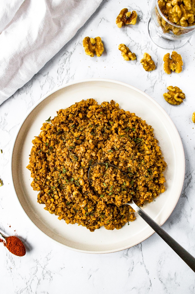 Overhead shot of sausage-style walnut meat on a white plate with a spoon digging into it. The plate is sprinkled with minced fresh herbs. There is a white linen, scattered raw walnuts, and a small spoon with smoked paprika on it surrounding the plate.