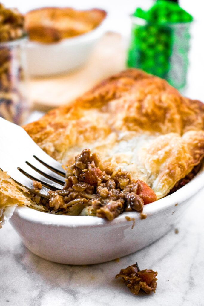 Head on photo of a fork digging into a savory vegetarian pie with puff pastry crust and walnut and mushroom filling. There is a jar of peas in the background along with another pie in a white ramekin.