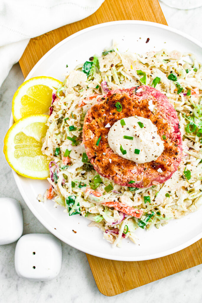 Overhead photo of a vegan salmon patty on a bed of tahini slaw with two lemon wedges to the side