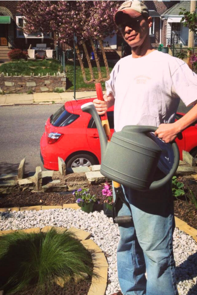A person with short hair and baseball hat holding a watering can and garden rake standing in a small city front yard garden